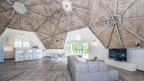 Een ruime loft met hoge plafonds, een keukenhoek, een eethoek en een woonkamer met een grote grijze bank.