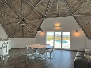 A spacious room with a high ceiling, a round table with chairs, and a view of a swimming pool through large windows.