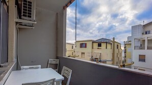 Un balcone con tavolo e sedie bianche, una finestra con vista sugli edifici vicini e un condizionatore d'aria a parete.