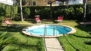 Piscina com deck ladrilhado, cercada por grama e palmeiras. Há duas espreguiçadeiras listradas em vermelho e branco.
