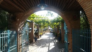 Um caminho de tijolos em arco com portões azuis, que leva a um jardim com plantas em vasos e um veículo branco ao fundo.