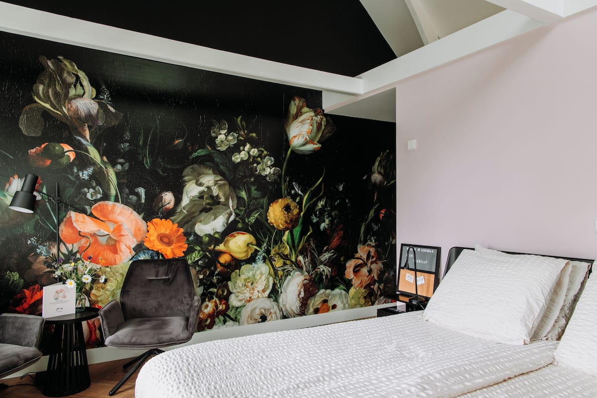 A bedroom with a large floral mural on the wall, a bed with white bedding, a grey armchair, and a small table with a vase of flowers.