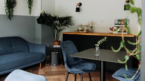 Una sala moderna con un sofá azul, una mesa de comedor con sillas, una planta en maceta y una pequeña área de cocina con encimera de madera y frascos.
