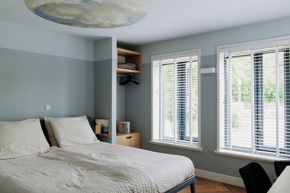 A bedroom with a bed, a nightstand, a dresser, a closet, and two windows with blinds.