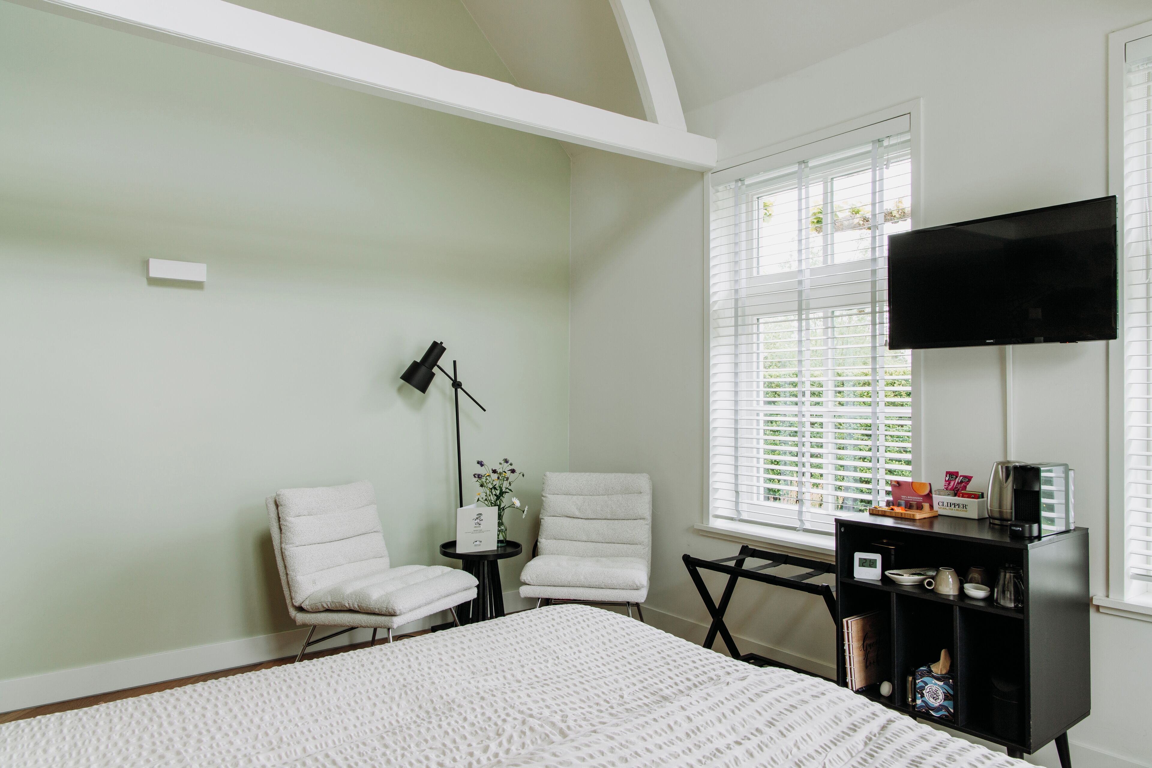 A bedroom with a bed, two chairs, a small table, a TV, and a window with blinds.