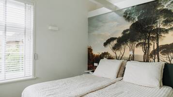 A bedroom with a bed, white bedding, a painting of trees, and a window with blinds.