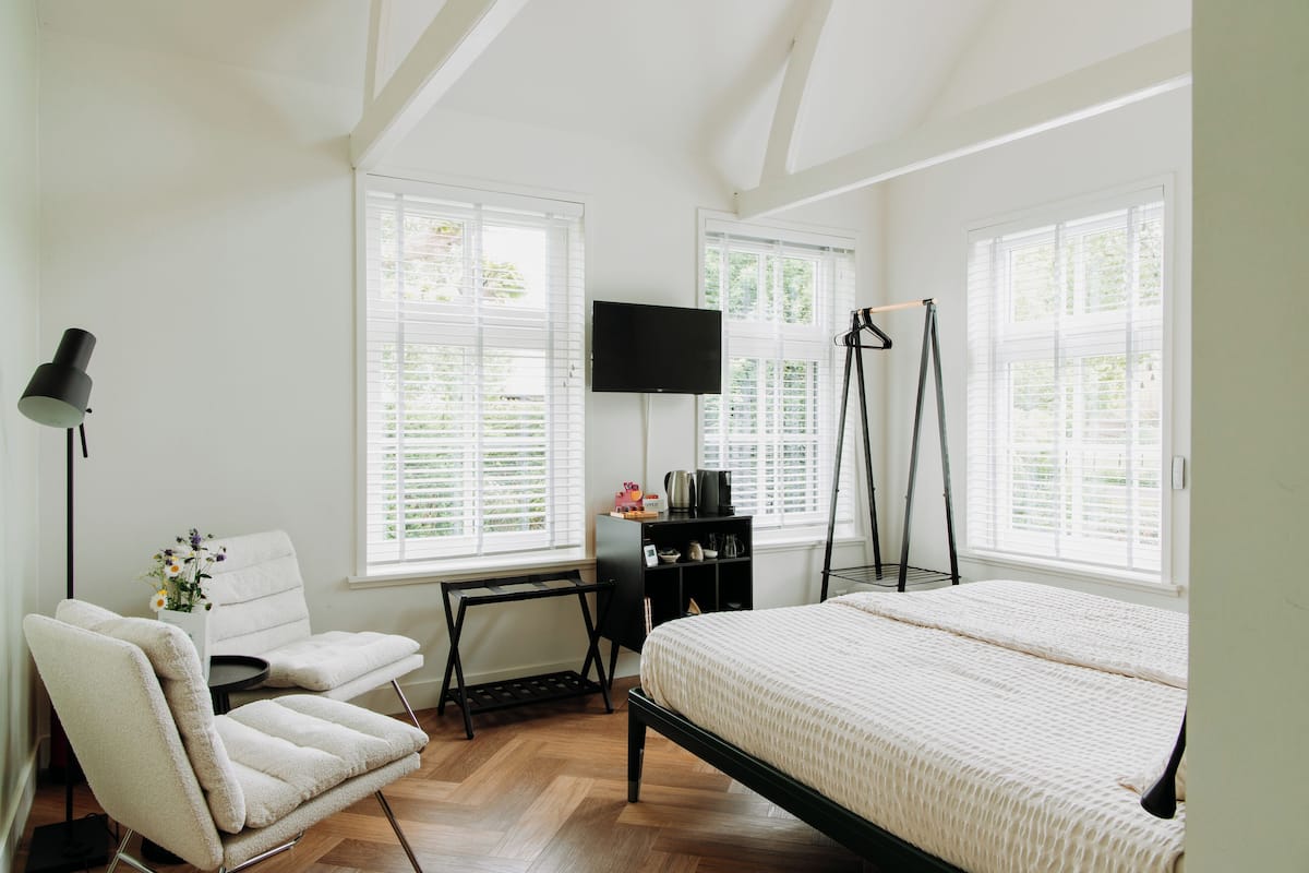 A bedroom with a bed, a chair, a desk, a TV, and large windows.