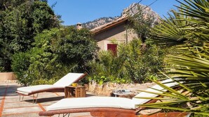 Un espacio al aire libre iluminado por el sol con sillones de descanso y una pequeña mesa, rodeado de exuberante vegetación y un edificio rústico.