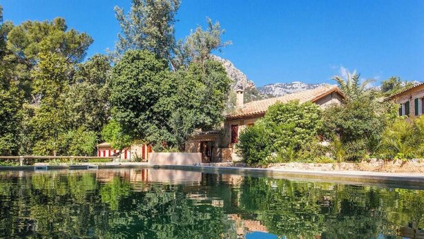 Un área de piscina tranquila con el reflejo de árboles y edificios, rodeada de exuberante vegetación y un paisaje montañoso.
