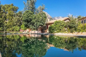 Uma área de piscina tranquila com reflexo de árvores e edifícios, rodeada por vegetação exuberante e uma paisagem montanhosa.