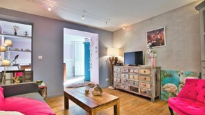 A living room with a wooden coffee table, a pink sofa, a red armchair, a TV, and a wall with a painting.