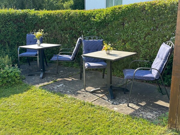 An outdoor seating area with metal chairs and tables, surrounded by a hedge and grass.