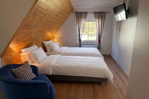 A bedroom with two beds, a wooden sloped ceiling, a TV mounted on the wall, and a window with curtains.
