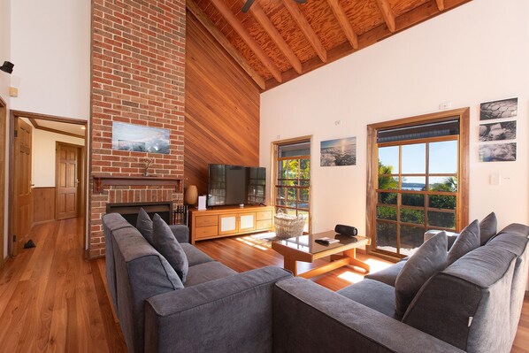 A spacious living room with a brick fireplace, wooden ceiling beams, and large windows offering a view of the outdoors.