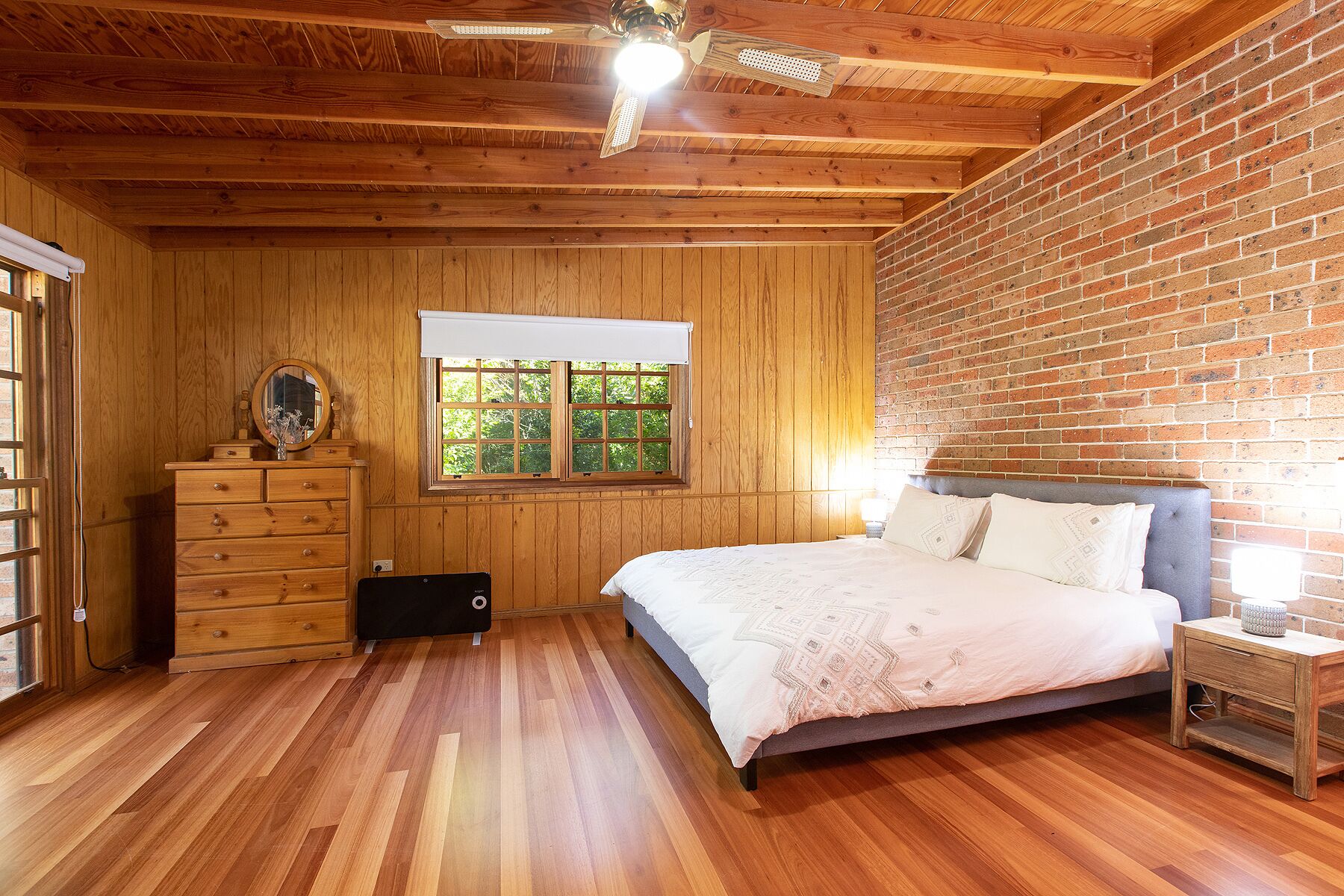 A bedroom with wooden walls, a brick accent wall, a bed with a white bedspread, a dresser, a mirror, and a window with a view of greenery.
