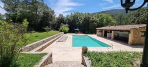 Une piscine rectangulaire bordée d’une terrasse en pierre, entourée d’un mur de pierre et d’une végétation luxuriante. Des chaises longues sont disposées le long du bord de la piscine, et un bâtiment en pierre avec un toit en tuiles est visible au fond.