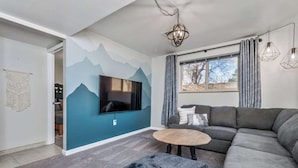 A modern living room with a large flat-screen TV, a gray sectional sofa, a round coffee table, and a mountain-themed wall mural.