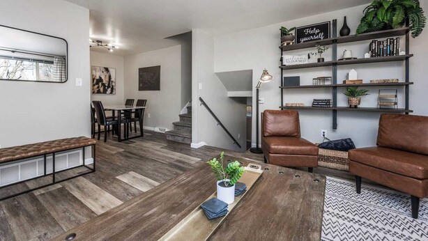 A modern living room with a brown leather sofa, a coffee table, and a bookshelf. There is a dining area with a table and chairs, and a staircase leading to another level.