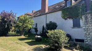 Une maison traditionnelle avec un mur en pierre, une cheminée en briques et un jardin agrémenté d’une brouette et d’arbustes.