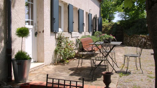 Un bâtiment doté d’une façade en pierre, d’une porte et d’une fenêtre avec des volets. On y trouve une terrasse avec une table, des chaises et des plantes en pot.
