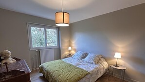 Une chambre à coucher avec un lit, deux lampes de chevet, une fenêtre, un radiateur et une commode en bois.