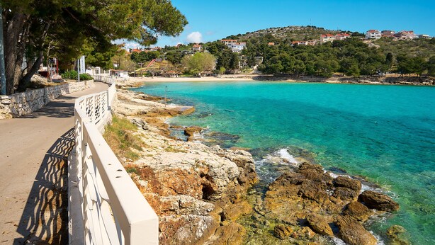 A coastal road with a stone railing, clear turquoise sea, rocky shoreline, and buildings on a hillside in the background. - Sun Spalato Villa Cikas (Murter-Kornati)