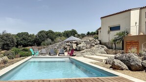 Une piscine rectangulaire avec une terrasse en bois au premier plan, entourée de rochers et de quelques chaises longues.