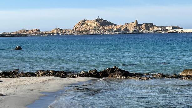 Une plage à l’eau claire, bordée d’affleurements rocheux, et une colline lointaine surmontée d’un phare.