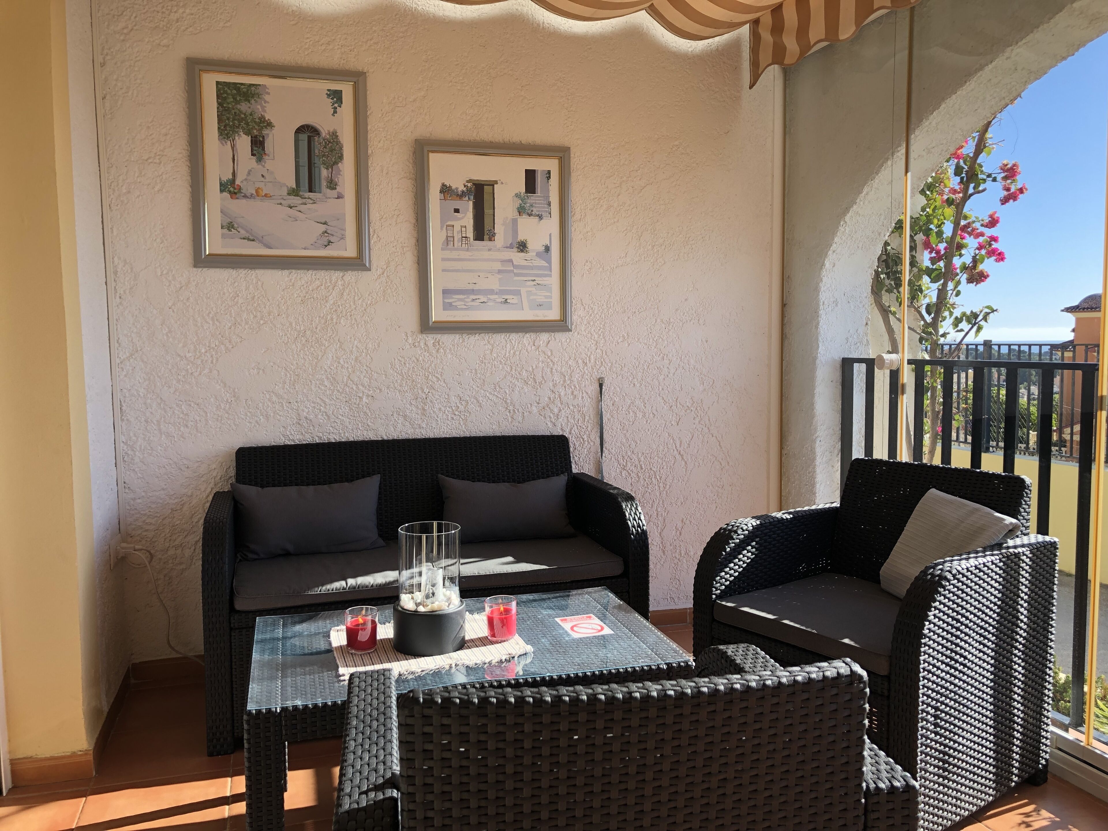 Un balcon agrémenté de meubles en osier, de deux œuvres d’art encadrées et d’une table en verre avec des bougies.