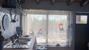 Una cocina con un lavamanos, estufa, ventana con cortina con motivos de gallinas y una puerta con mosquitero.