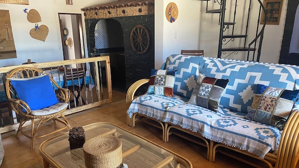 Una sala de estar con muebles de mimbre, una mesa de centro de vidrio, un sofá estampado y una chimenea de piedra.