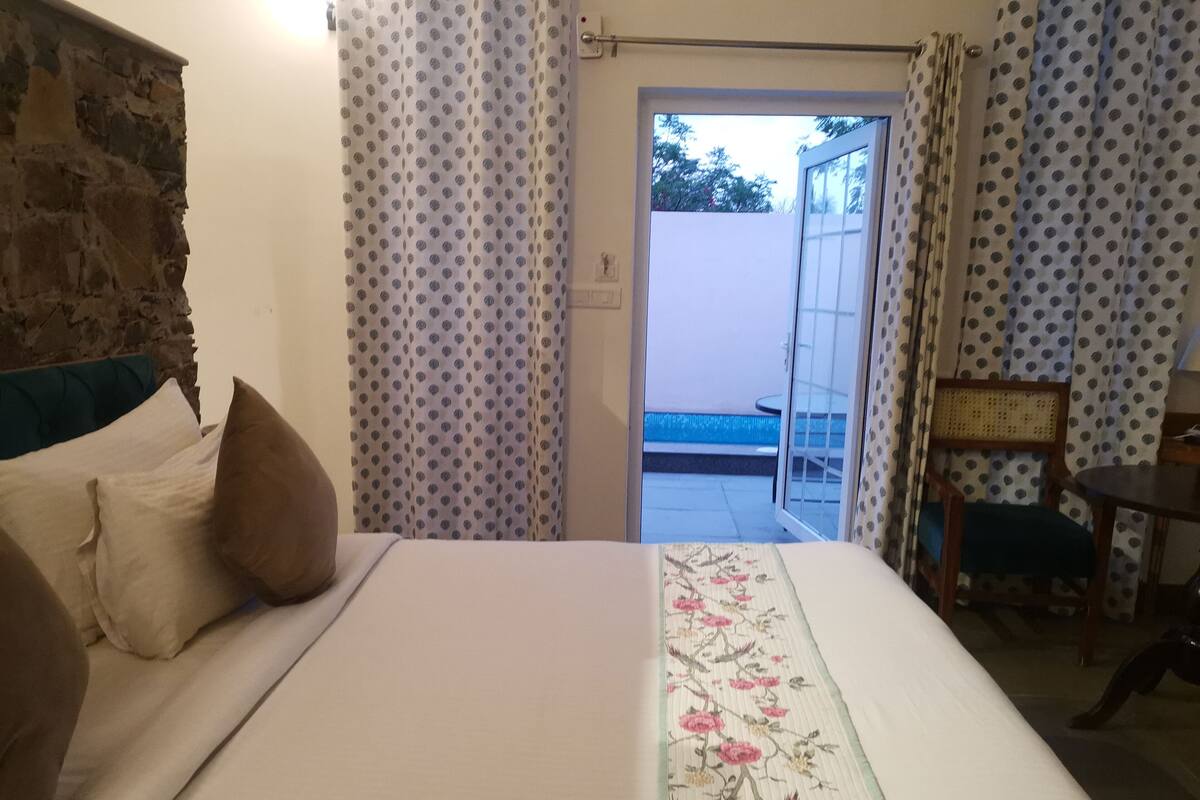 Ein Schlafzimmer mit Bett, Kissen, einem Holz-Kopfende, einem kleinen Tisch, einem Sessel und Blick auf einen Außenbereich mit einem Pool.