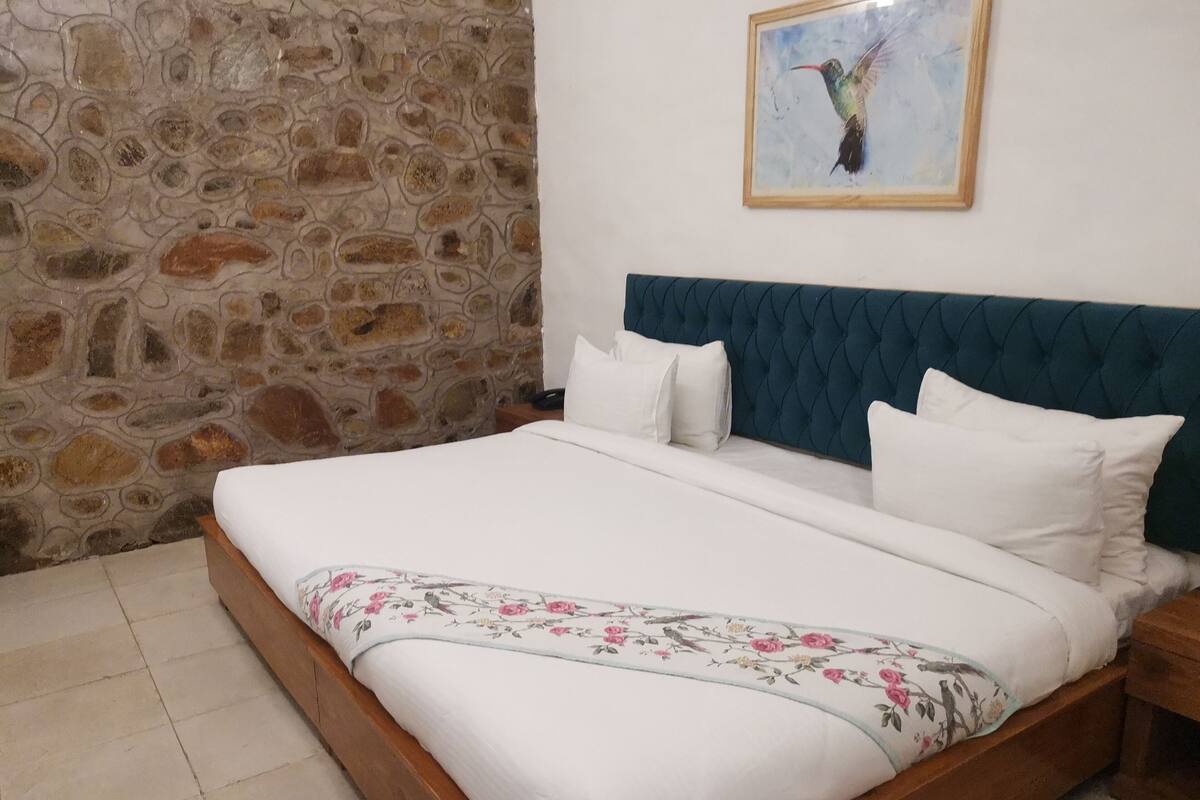 Ein Schlafzimmer mit Stehwänden, einem Bett mit weißer Bettwäsche und Blumenmuster, einem blauen, gepolsterten Kopfteil und einem Bilderrahmen mit einem Kolibri.
