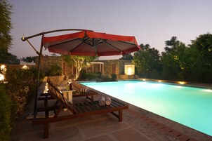 Zona junto a la piscina con sillones de descanso y una sombrilla roja, rodeada de muros de piedra y vegetación.