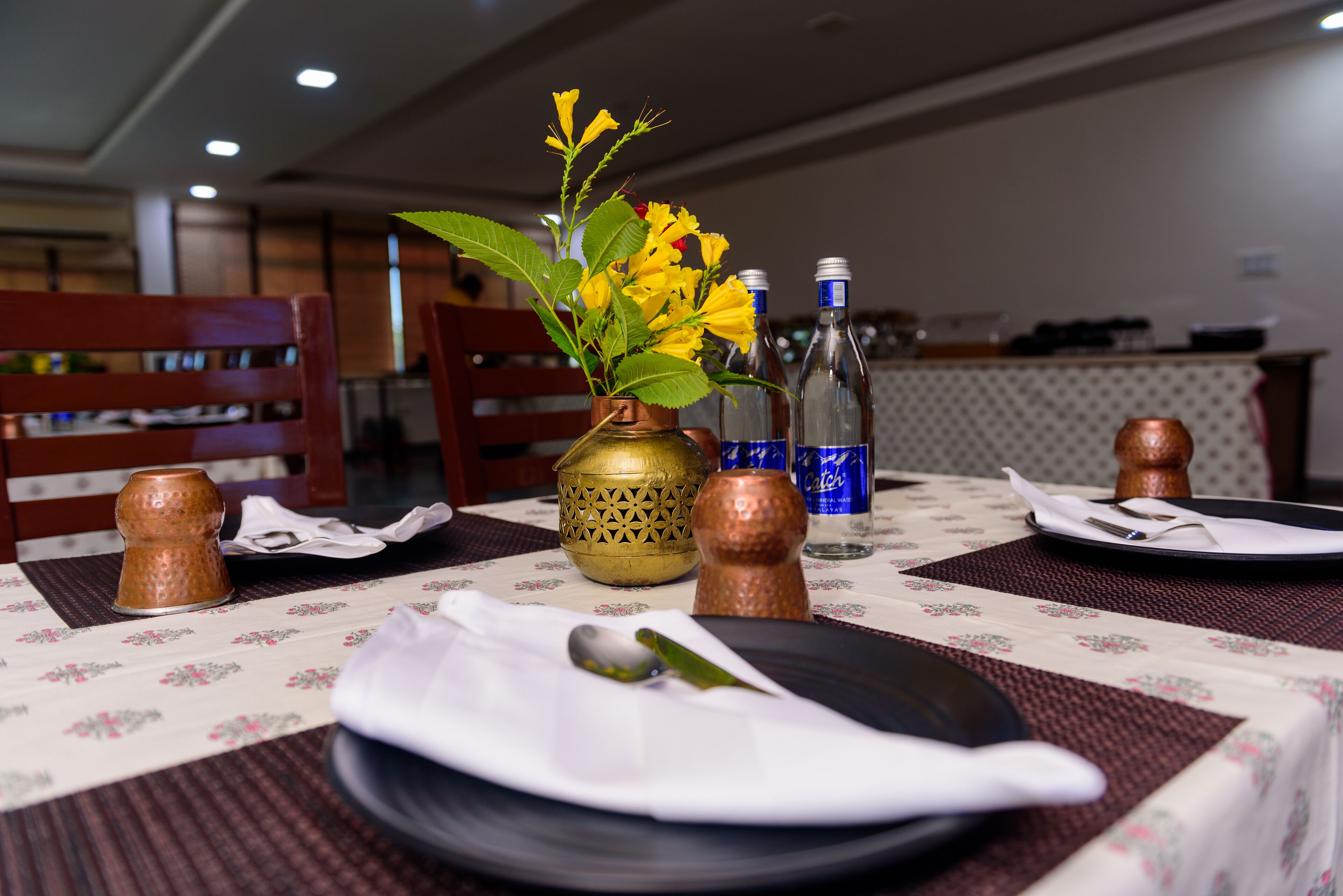 A dining table with a floral tablecloth, a vase with yellow flowers, and copper salt and pepper shakers.