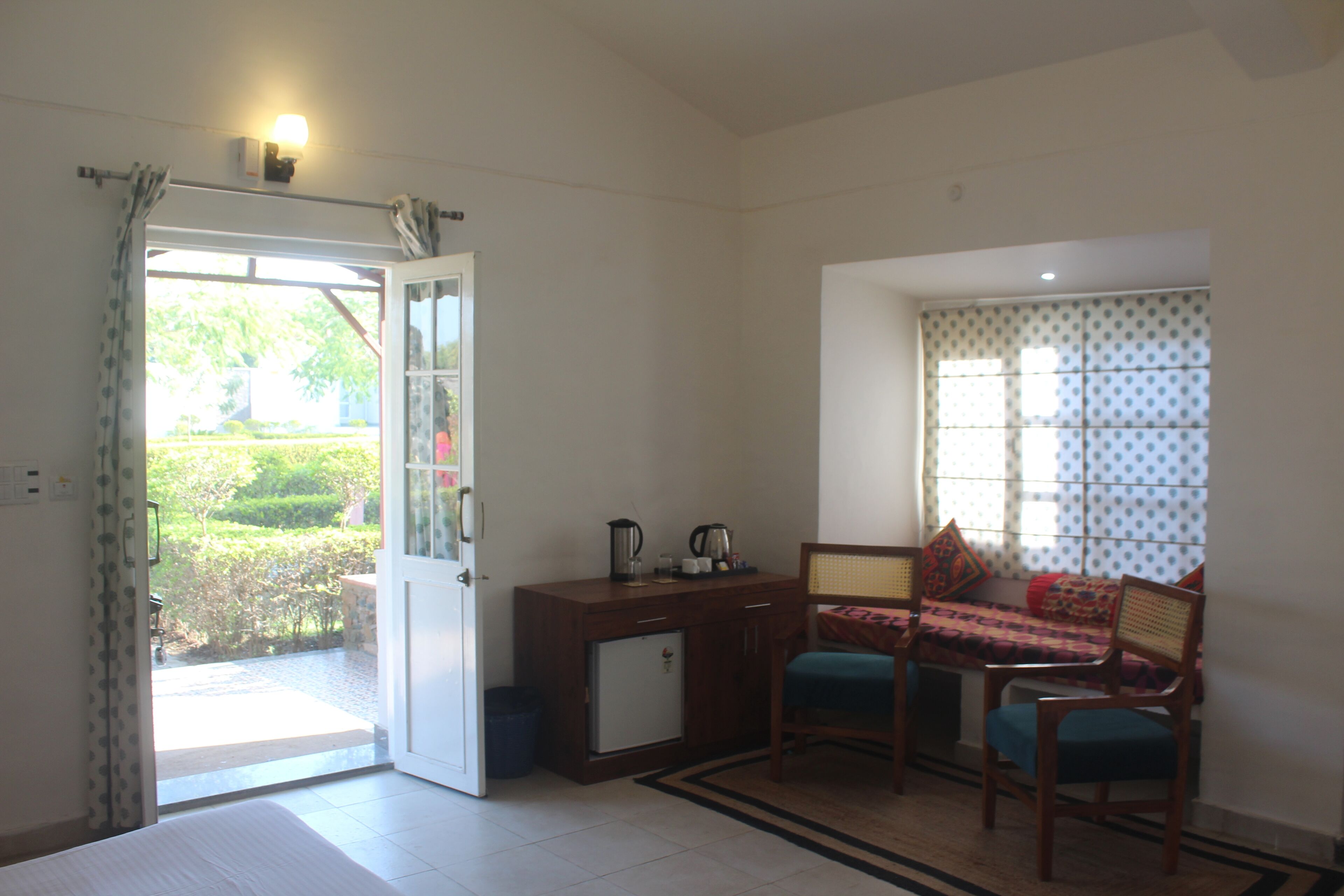 A room with a bed, a small desk with a kettle, a chair, and a window with a view of greenery.