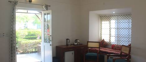 A room with a bed, a small desk with a kettle, a chair, and a window with a view of greenery.