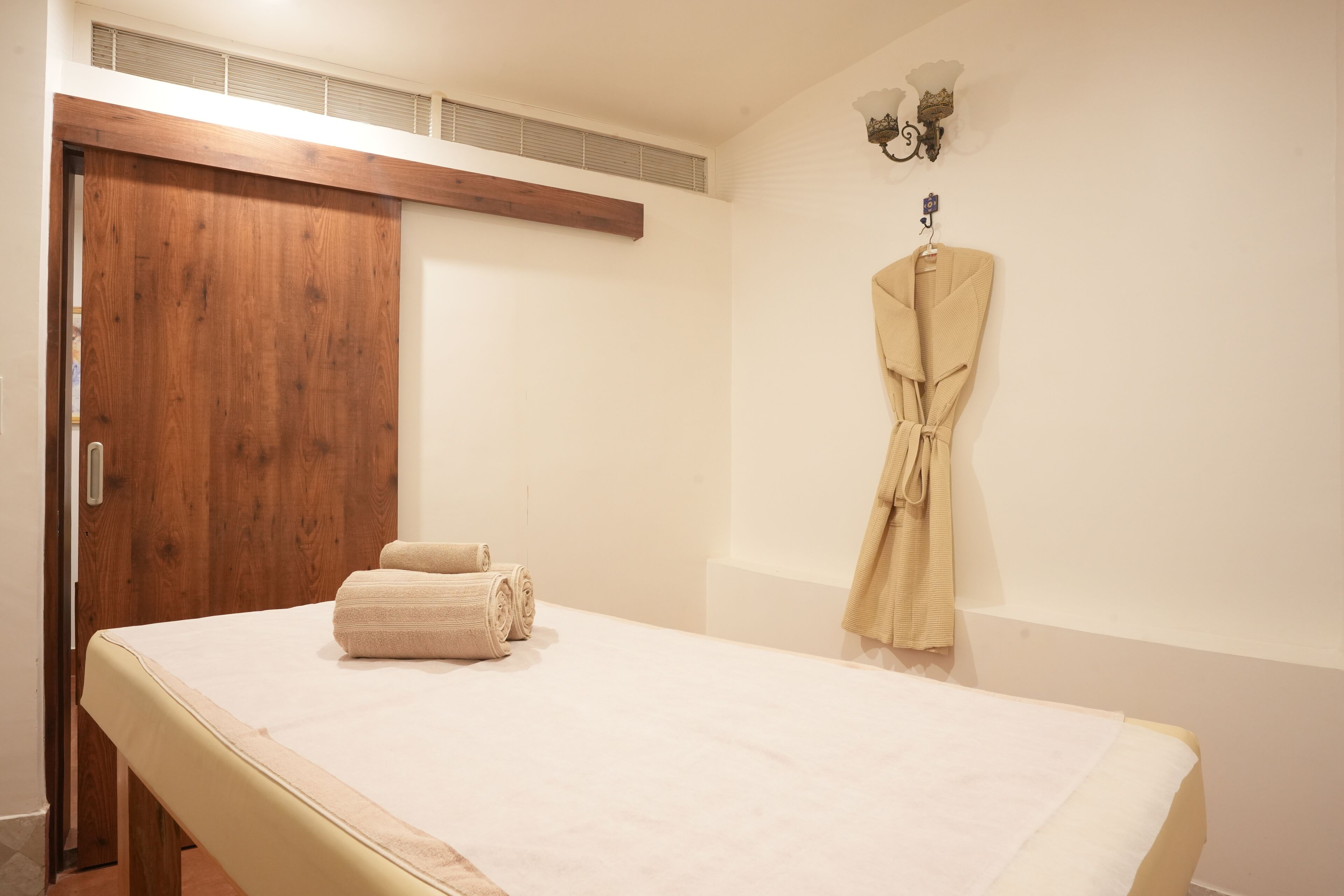 A room with a wooden door, a bed with a white mattress, and a beige robe hanging on the wall.