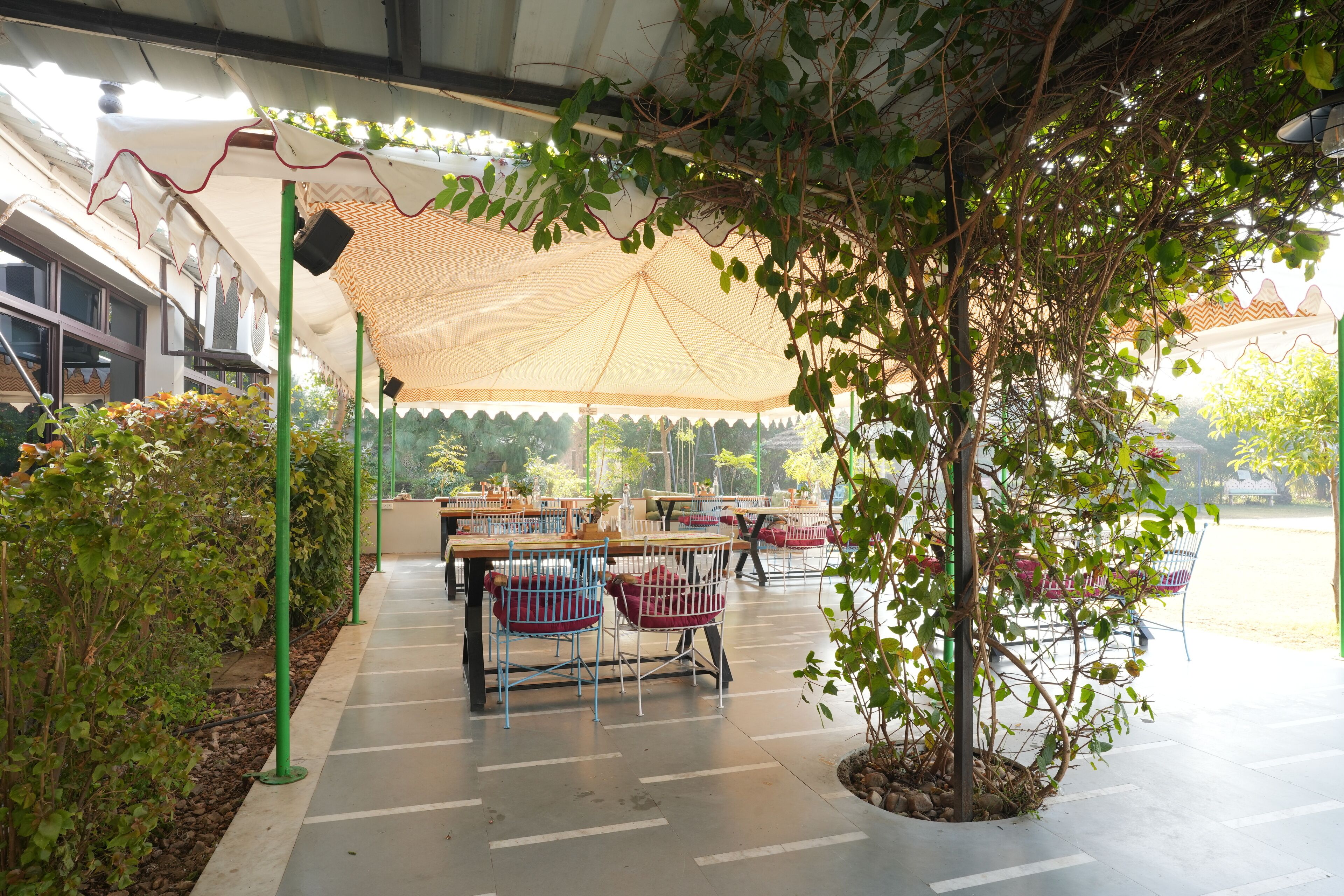 An outdoor seating area with tables and chairs under a shaded canopy, surrounded by greenery.