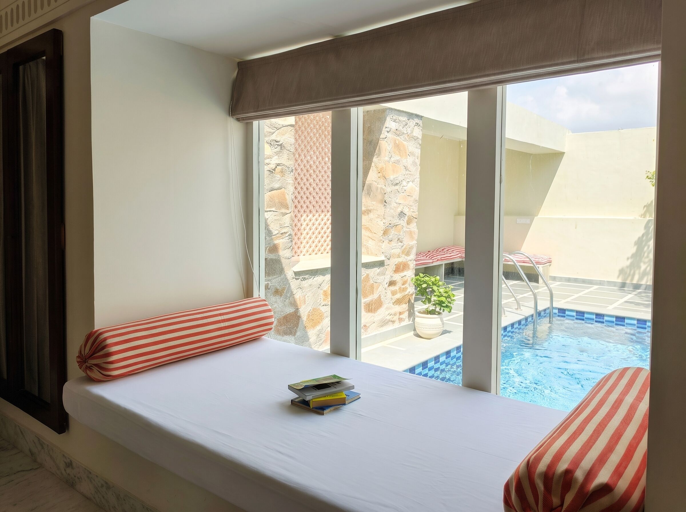 A hotel room with a large window, a bed with striped pillows, a small table with books, and a view of an outdoor swimming pool.