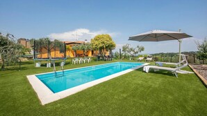 A swimming pool with a white deck, surrounded by green grass, and a patio area with chairs and an umbrella. - Villa Teresa Apt Lake Garda View 2BR + Pool (Soiano del Lago)