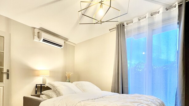 A modern bedroom with a large bed, a bedside table with a lamp, an air conditioning unit, and a view of the outdoors through a window with curtains. - Charming T2 with terrace and private parking (Champagne-au-Mont-d'Or)
