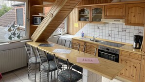 Une cuisine avec des meubles en bois, une table à manger avec des chaises, un four encastré et une fenêtre agrémentée d’objets décoratifs.