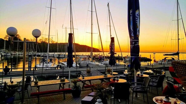 Un port de plaisance avec des bateaux amarrés, un coucher de soleil en arrière-plan et des espaces de détente en plein air.