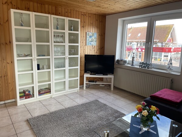 A living room with a TV, a bookshelf, a window with a view of buildings, and a table with a vase of flowers. - Odin's little sea view (Langballig)