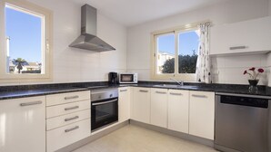 A modern kitchen with white cabinets, a stainless steel dishwasher, a built-in oven, a microwave, and a window with curtains.