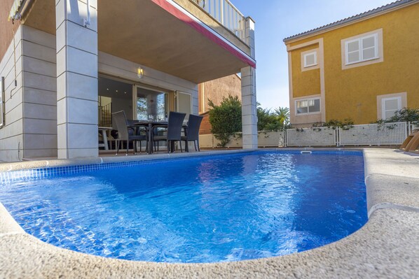 A swimming pool with a covered patio area, a dining table, and chairs.