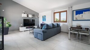 Una sala moderna con un sofá gris, un televisor de pantalla plana, una mesa de comedor con sillas y un ventanal con cortinas.