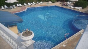A rectangular swimming pool with clear blue water, surrounded by white lounge chairs and umbrellas, with a balustrade and a flower pot in the foreground. - Dahlia Garden (Golden Sands)