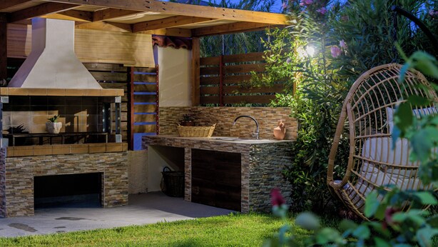 An outdoor kitchen with a stone fireplace, a sink, and a wooden pergola overhead. - Diogenis House (Zakynthos)
